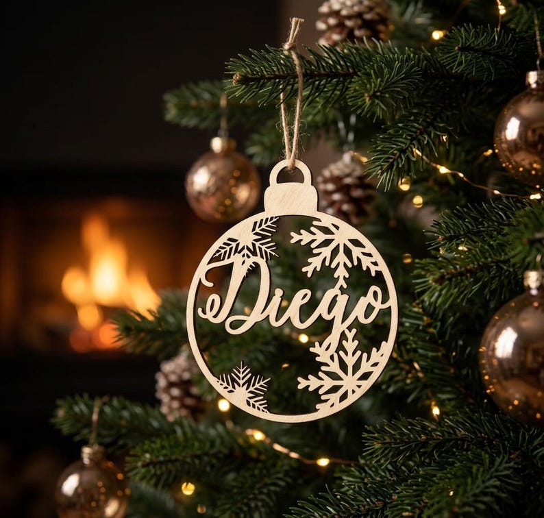 May include: A circular light-colored wooden Christmas ornament featuring a cutout design of snowflakes and the name Diego in a cursive script font. The ornament is suspended by a thin twine string from a branch of a green pine tree.
