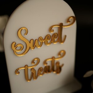 May include: White sign with a gold cursive inscription that reads "Sweet Treats".