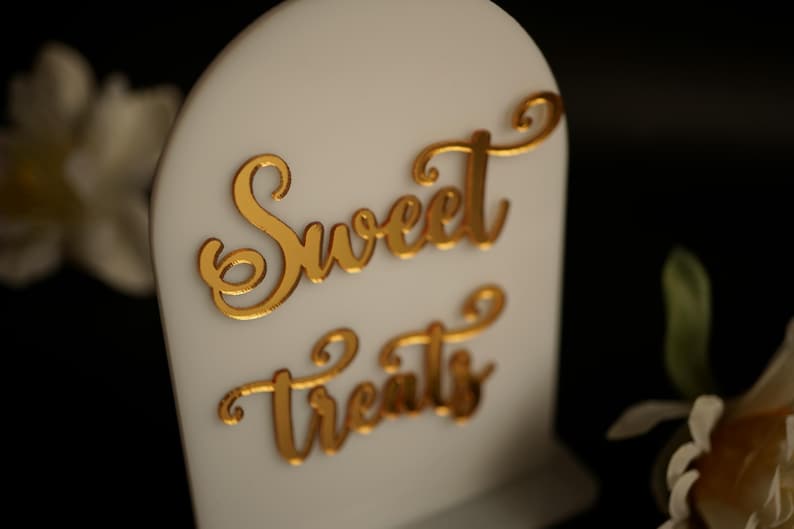 May include: White sign with a gold cursive inscription that reads "Sweet Treats".