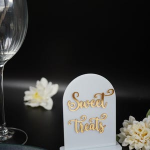 May include: White acrylic sign with a gold inscription that reads "Sweet Treats". The sign is on a black surface with a white flower and a gold fork and spoon.