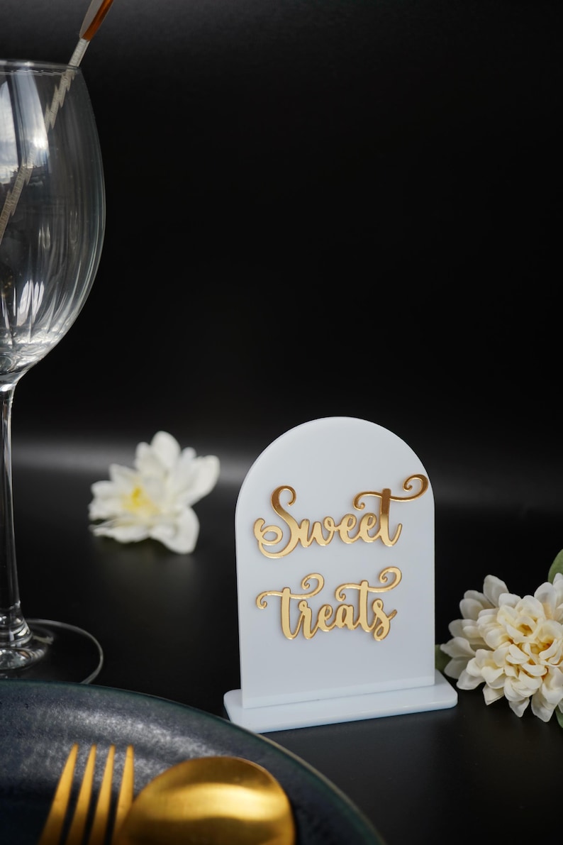 May include: White acrylic sign with a gold inscription that reads "Sweet Treats". The sign is on a black surface with a white flower and a gold fork and spoon.
