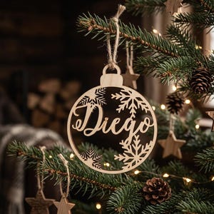 May include: A circular light-colored wooden Christmas ornament featuring the name Diego in a cursive script, surrounded by intricate laser-cut snowflake patterns. The ornament hangs from a natural jute twine loop on a green pine tree branch.