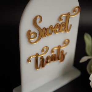 May include: White acrylic sign with a gold arch top and a gold cursive inscription that reads "Sweet Treats".