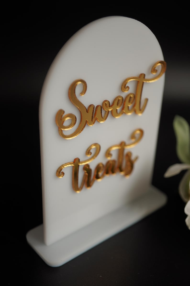 May include: White acrylic sign with a gold arch top and a gold cursive inscription that reads "Sweet Treats".