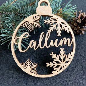 May include: A circular light-colored wooden ornament featuring the name Callum in a cursive script font. The design includes cut-out snowflake patterns along the top and bottom edges and a small loop at the top for hanging purposes.