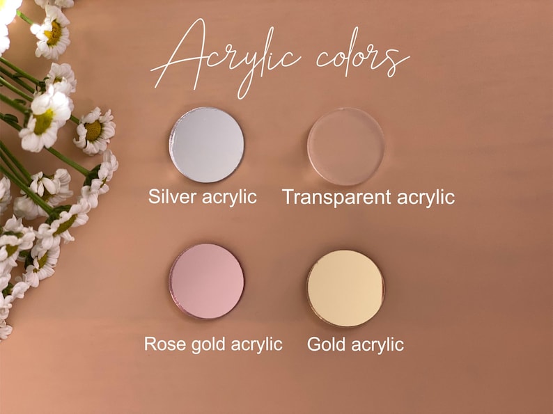 May include: Four circular acrylic discs arranged in a grid, labeled as silver, transparent, rose gold, and gold acrylic. The text "Acrylic colors" appears at the top. White daisy flowers are positioned along the left side of the display.