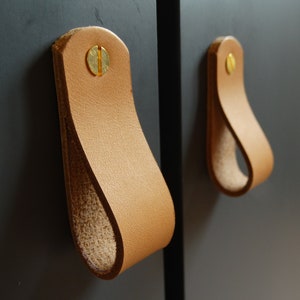 Peut inclure: Deux poignées de tiroir en cuir marron avec des vis en laiton montées sur une surface gris foncé.