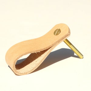 Könnte beinhalten: Ein hellbrauner Leder-Schrankgriff mit einer Messing-Schraube. Der Griff ist schlauchartig geformt und hat eine glatte, leicht strukturierte Oberfläche.