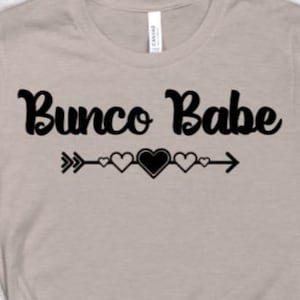 May include: A light tan t-shirt with the text "Bunco Babe" in a black script font. Below the text is a black arrow with three hearts. The shirt is made of a soft material and has short sleeves.