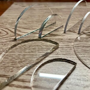 May include: A collection of clear, curved, and semi-circular glass pieces with silver edges are arranged on a wooden surface. The shapes vary in size and are positioned at different angles, creating a display of light and shadow.