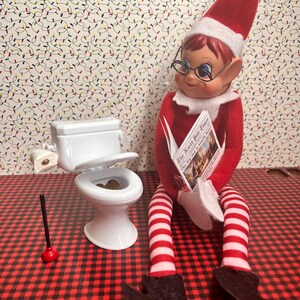 May include: A red and white elf doll wearing glasses and a red hat, sitting on a miniature toilet. The elf is reading a newspaper titled "The North Pole Times". The background features a wallpaper with Christmas lights.