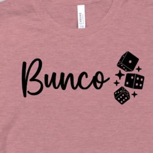 May include: Dusty rose t-shirt with the word "Bunco" in a cursive font and three black dice with white dots. The dice are accompanied by small star graphics.