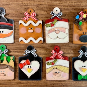 May include: A collection of eight wooden Christmas ornaments. Each ornament is a rectangular shape with a handle and a decorative bow. The ornaments feature festive designs like a snowman, gingerbread man, Santa, and a reindeer.