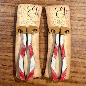 May include: Two wooden Christmas ornaments shaped like elf legs. The ornaments are painted with red and white stripes and have brown elf shoes. The text "It's Elf Season" is written on the top of each ornament.