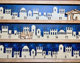 May include: A wooden advent calendar with a blue starry sky background. The calendar features 24 numbered doors, each with a unique cutout design. The top row features a nativity scene with a stable and figures of Mary, Joseph, and the baby Jesus.