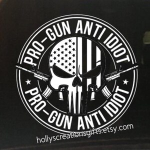 May include: A black and white sticker featuring a skull design with an American flag motif. The text "PRO-GUN ANTI IDIOT" encircles the skull, with two rifles and the Etsy shop URL. The design is bold and graphic.
