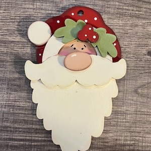 May include: Wooden Santa Claus face ornament. The ornament features a red hat with white polka dots, a white beard, and a pink nose. Green holly leaves and red berries adorn the hat. The ornament has a hole at the top for hanging.