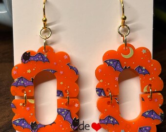 May include: Orange Halloween-themed earrings with a scalloped edge design. Each earring features a bat and star pattern with purple and black accents. The earrings have gold-colored hooks.