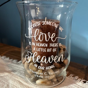 May include: A clear glass vase with a brown candle inside. The vase is filled with small stones and has a white inscription that reads "Because someone we love is in heaven there is a little bit of heaven in our home Thomas C. Louer 1948-2021".