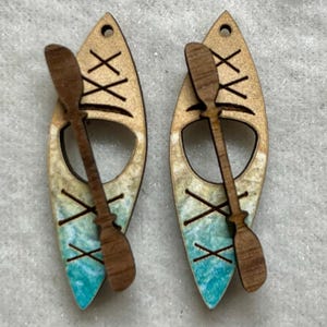 May include: A pair of wooden earrings shaped like kayaks with paddles. The kayaks are painted with a blue and white water design and have a brown wood grain finish.
