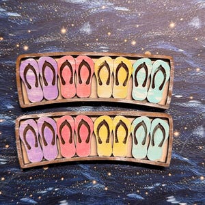 May include: Two curved wooden trays, each holding eight small, flat flip-flop cutouts arranged in pairs. The flip-flops are painted in purple, pink, yellow, and light blue, featuring dark brown straps. The trays have a natural wood finish.