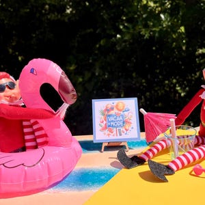 May include: Two elves on a beach scene. One elf is riding a pink inflatable flamingo float. The other elf is sitting on a yellow beach towel with a red and white striped towel, a glass of water, and a pink umbrella. A sign reads "Vacay Mode".