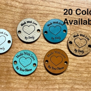 May include: Six circular tags in various colors including white, teal, light blue, brown, and cork, each featuring two side holes. The tags are engraved with a heart icon and the text Made With Love followed by By Emily or By Your Name.