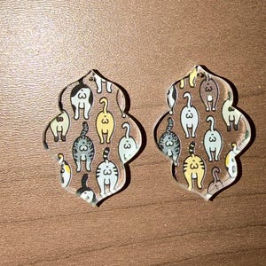 May include: Two clear acrylic earrings with a repeating pattern of cat bottoms in various colors. The earrings have a unique quatrefoil shape and are designed to be worn as a pair. The cat bottoms are in shades of gray, yellow, and white.