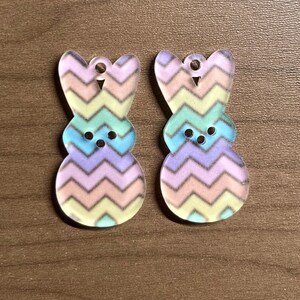 May include: Two Easter bunny-shaped acrylic charms. Each charm features a pastel chevron pattern in shades of yellow, green, blue, and purple. The charms have small holes at the top for hanging and two small circles for eyes.