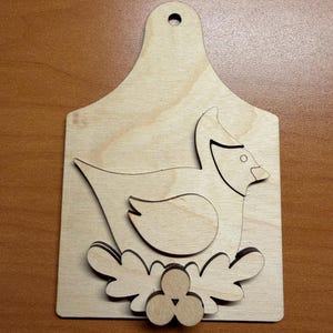 May include: A light-colored wooden plaque shaped like a cutting board with a hole at the top. It features a layered, laser-cut design of a bird sitting in a nest with three small circular berries at the base, all made from natural wood.