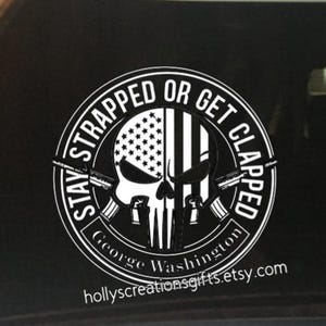 May include: A circular white sticker with a skull design, half of which is the American flag. The text "STAY STRAPPED OR GET CLAPPED" surrounds the skull. The text "George Washington" and a website address are also included.