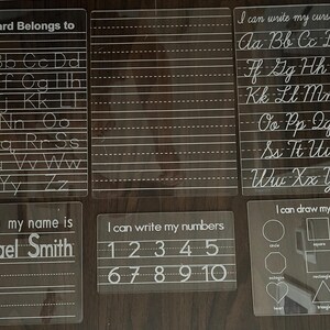 May include: Six clear plastic dry-erase boards with white writing. The boards include the alphabet, cursive letters, numbers, and shapes. One board says "This Board Belongs to" and has a space for a name. Another board says "Hello my name is" and has a space for a name.