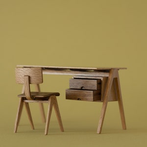 May include: A miniature wooden desk and chair set. The desk has two drawers and angled legs. The chair has a curved back and seat. The set is made of light-colored wood and is set against a solid yellow-green background.