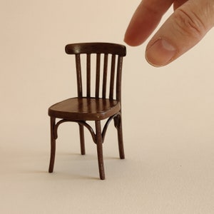 May include: A miniature wooden chair with a dark brown finish. The chair has a curved back with vertical slats and a rounded seat. The legs are also curved, and the chair appears to be a small-scale replica.