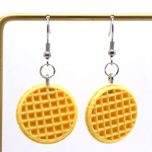 May include: A pair of novelty earrings featuring a yellow waffle design. The earrings are round with a grid pattern and are suspended from silver-colored hooks. The earrings are displayed against a white background.