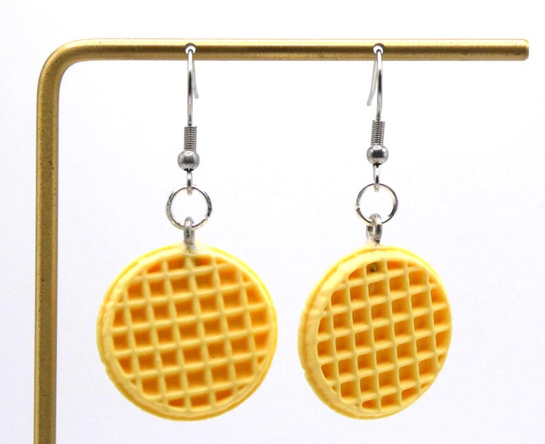 May include: A pair of novelty earrings with a yellow waffle design. The earrings are round with a grid pattern and are suspended from silver-coloured hooks. The earrings are displayed against a white background.