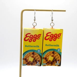 May include: A pair of novelty earrings featuring miniature Eggo Buttermilk waffle boxes. The boxes are yellow with red and blue text, and a picture of a waffle with butter. The earrings hang from silver-colored hooks.