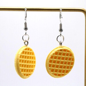 May include: A pair of novelty earrings shaped like waffles. The earrings are yellow with a grid pattern, hanging from silver-coloured hooks. The earrings are displayed against a white background.