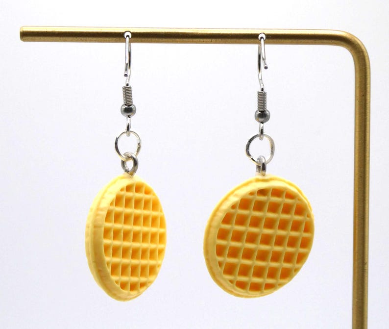 May include: A pair of novelty earrings shaped like waffles. The earrings are yellow with a grid pattern, hanging from silver-coloured hooks. The earrings are displayed against a white background.