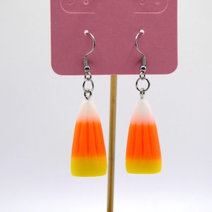 Pode incluir: Um par de brincos de doces de milho. Cada brinco apresenta um gancho prateado e um berloque em forma de doce de milho. Os berloques de doces de milho são brancos no topo, passando para laranja e terminando com amarelo na parte inferior.