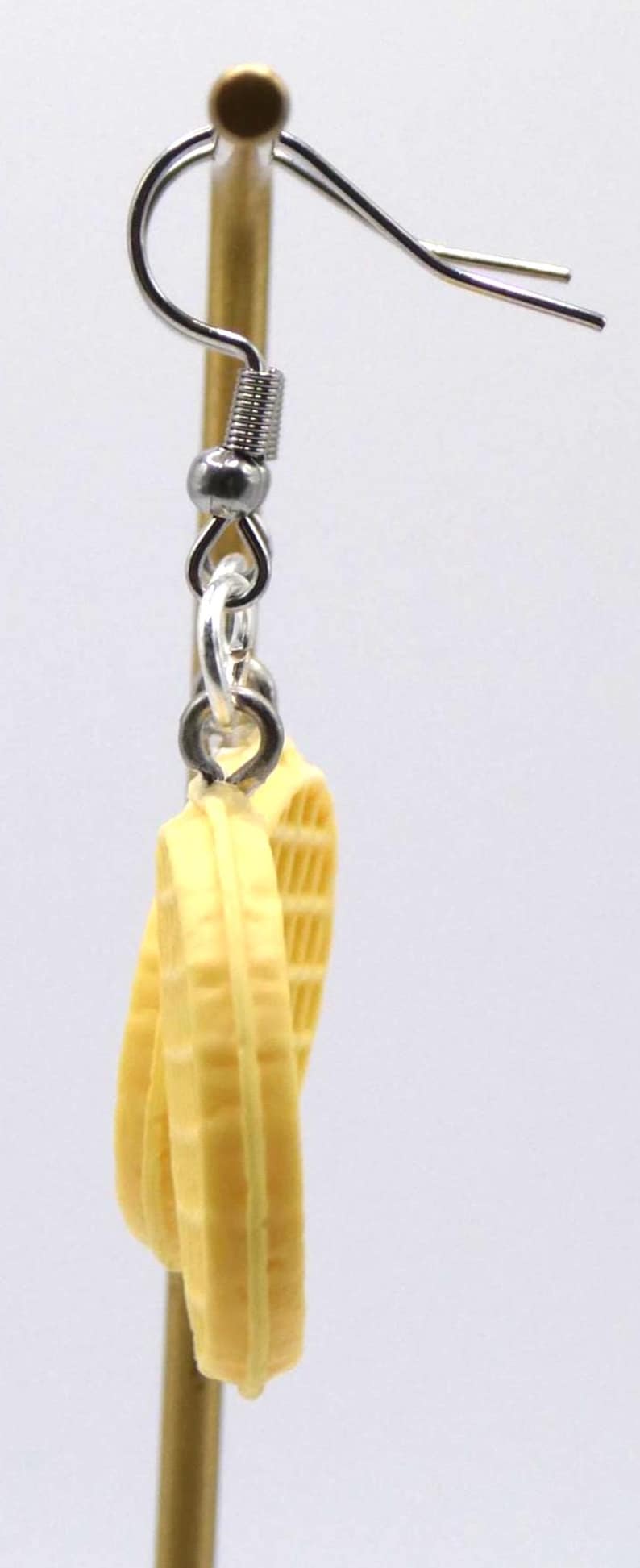 May include: A pair of novelty earrings with a waffle design. The earrings are light yellow and have a silver-coloured hook. The waffle earrings are a fun and unique accessory.