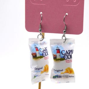 Può includere: Un paio di orecchini originali con mini sacchetti di patatine Cape Cod. I sacchetti mostrano una scena di faro e le parole "Cape Cod" e "Original Sea Salt". Gli orecchini sono appesi a ganci color argento.