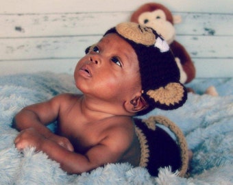 May include: A baby wearing a brown crocheted monkey hat with a tan face and ears. The baby is lying on a blue fuzzy blanket and looking up.