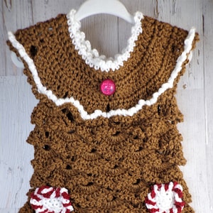 May include: A brown crocheted gingerbread house dress with white trim and candy cane accents. The dress has a pink button on the front.