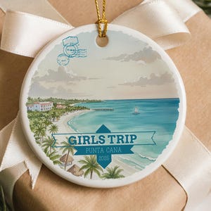 May include: A round ceramic ornament with a beach scene, the words "GIRLS TRIP PUNTA CANA 2025" are printed on the front. The ornament is on a wrapped gift box with a cream ribbon and pine needles.