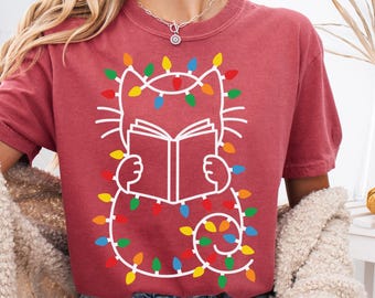 Op de afbeelding: Een bordeauxrood t-shirt met een witte omtrek van een kat die een boek leest, versierd met kleurrijke kerstverlichting. Het kattenontwerp staat centraal op het shirt. Het model draagt een spijkerbroekshort en een crèmekleurig vest.