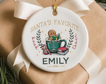 May include: White ceramic Christmas ornament with gold text that reads "Santa's Favorite Book Lover" and the name "Emily". The ornament features a gingerbread man in a mug, books, and candy canes. A gold string is attached.