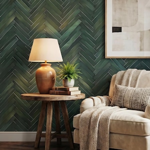 Op de afbeelding: Een crèmekleurige fauteuil met een textuurrijke plaid en een kussen met patroon, naast een ronde houten bijzettafel met een lamp, een kleine plant in pot en twee boeken, geplaatst tegen een donkergroene muur met visgraatmotief.