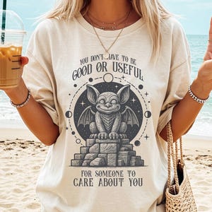 Könnte beinhalten: Beiges T-Shirt mit einem grauen Gargoyle-Design in einem Kreis und dem Text: "YOU DON'T HAVE TO BE GOOD OR USEFUL FOR SOMEONE TO CARE ABOUT YOU." Das T-Shirt wird am Strand getragen, mit einem Getränk in der Hand.