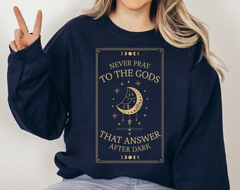 May include: A navy blue sweatshirt with a gold graphic print of a crescent moon and stars. The text on the graphic reads "Never pray to the gods that answer after dark".